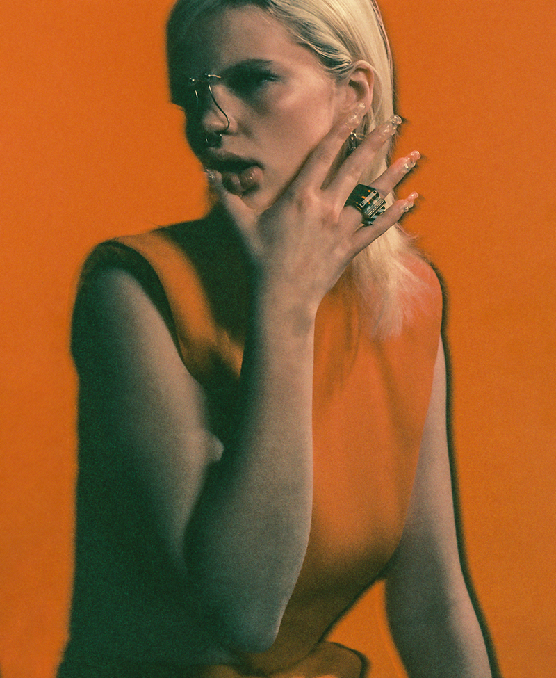 A woman with blond hair, wearing glasses and an orange sleeveless top, poses against a vibrant orange backdrop.
