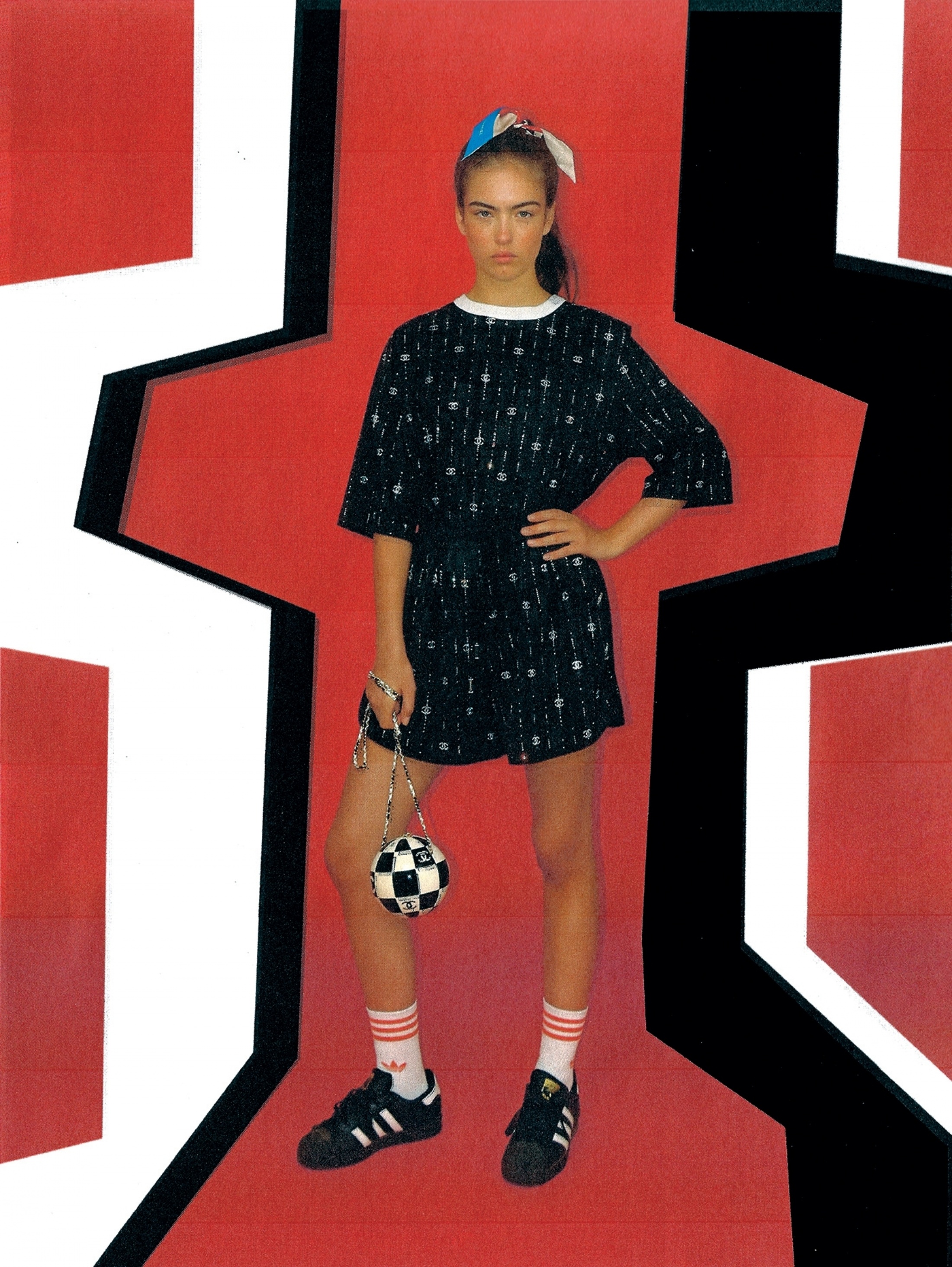 Young woman in patterned shirt and shorts holding soccer ball purse, posing in a geometric, vibrant red and black design.