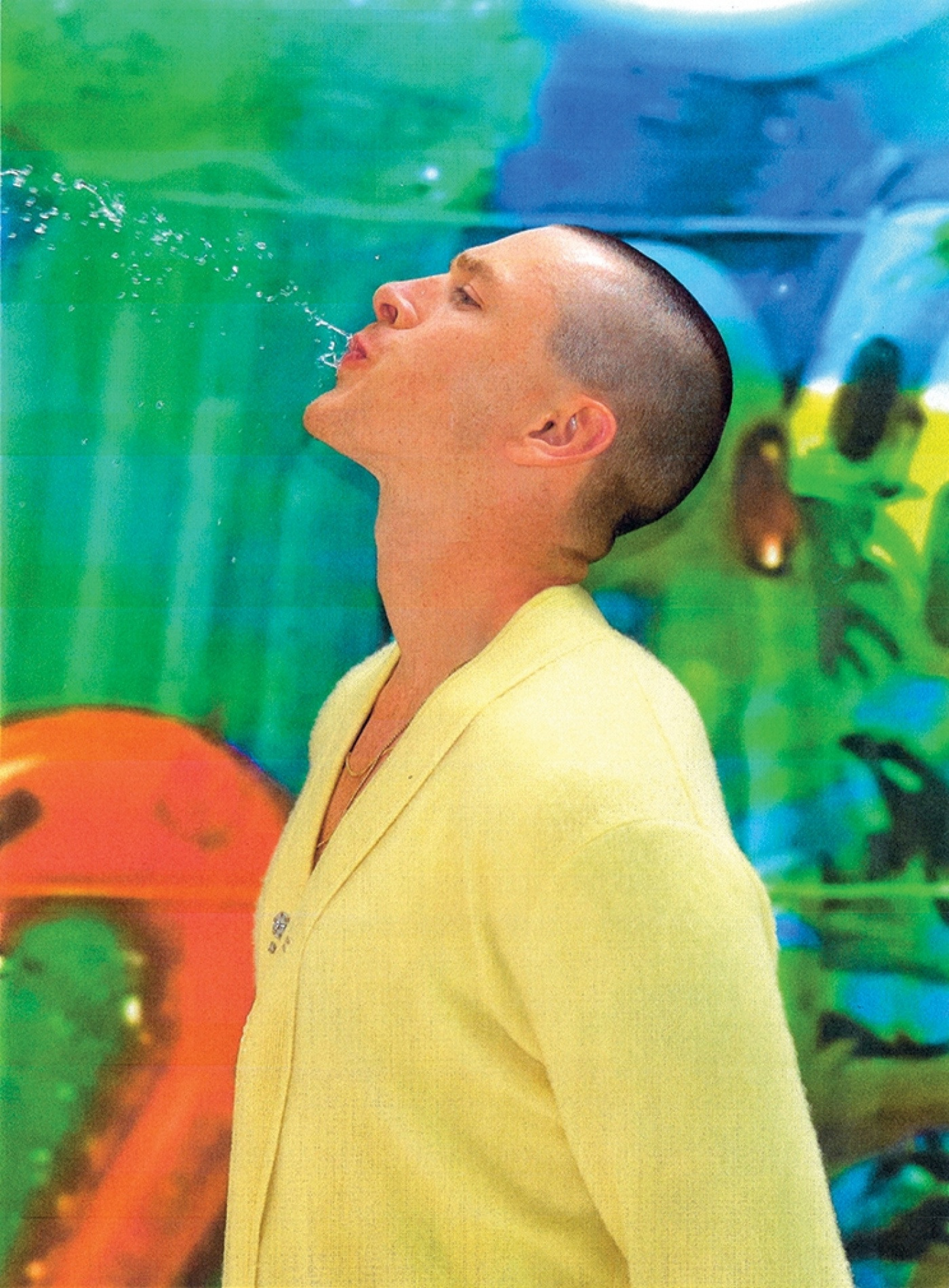 Young man in a yellow jacket spitting water from his mouth against an abstract art background in a studio setting, looking artistic.