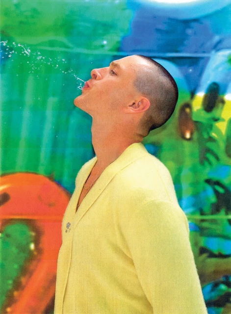 Young man in a yellow jacket spitting water from his mouth against an abstract art background in a studio setting, looking artistic.