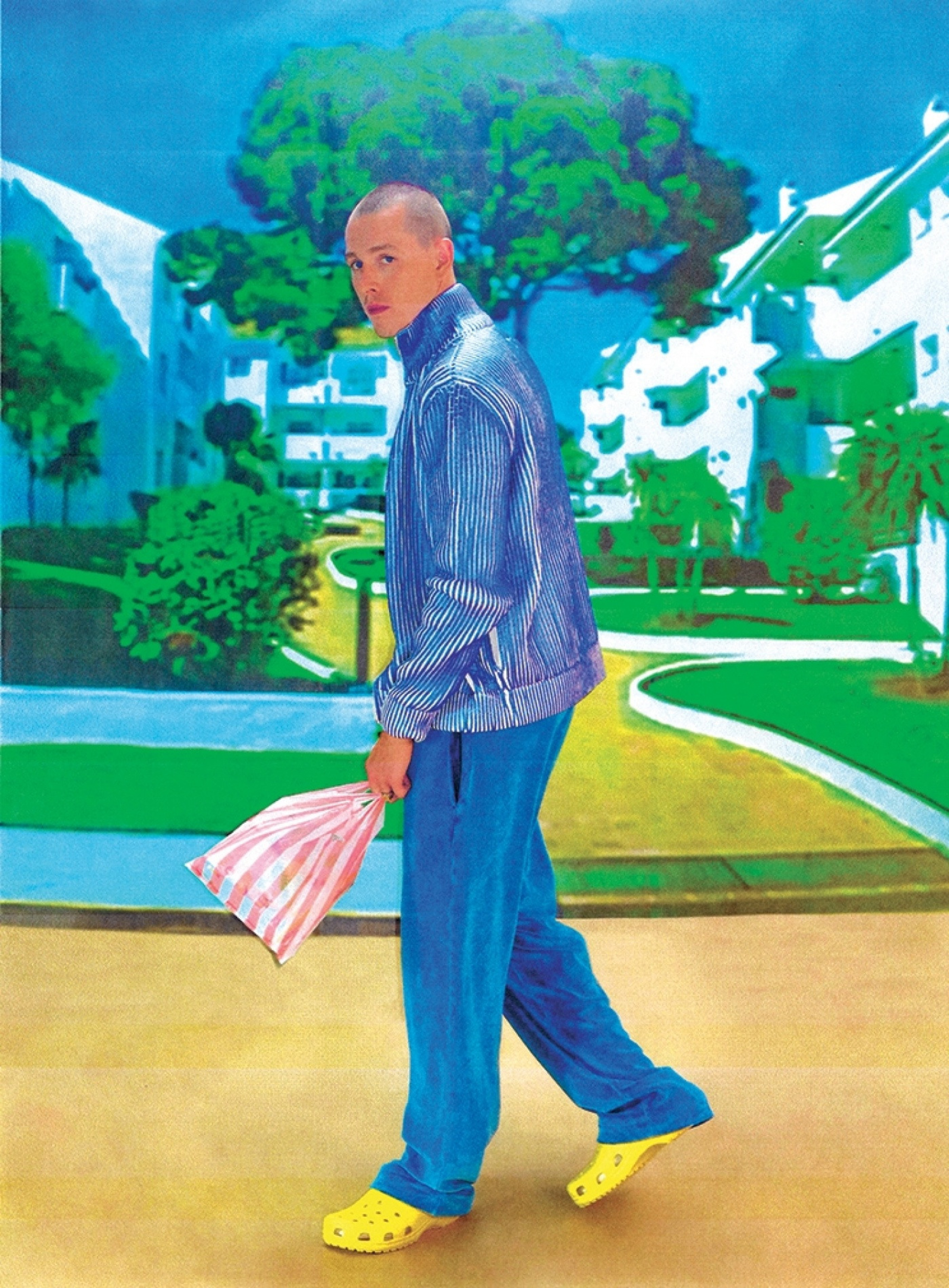 Man in blue striped jacket and pants walks in a neighborhood with yellow shoes and a striped bag with blue sky and white buildings.