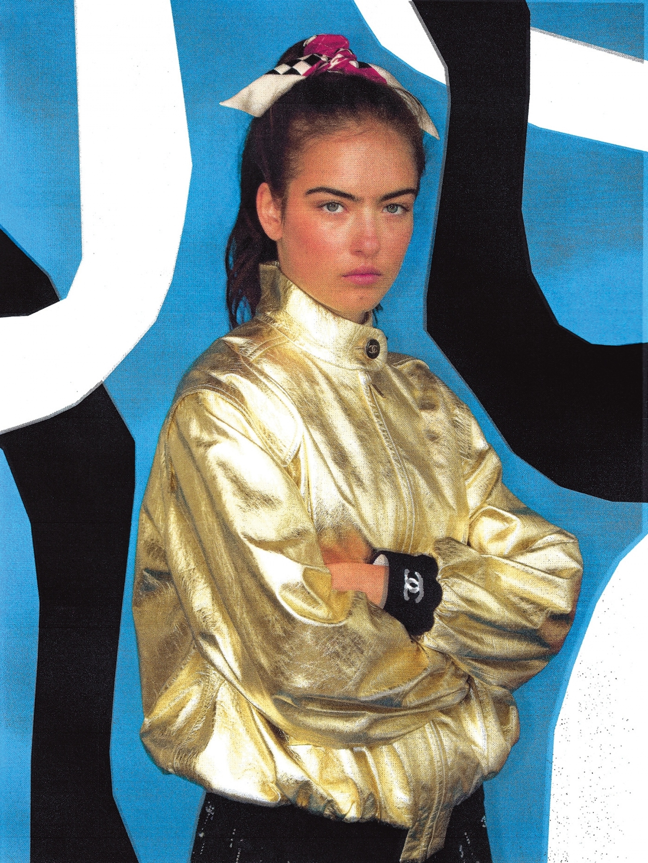 A young woman with her arms crossed wears a gold jacket, a scarf in her hair and black gloves against an abstract blue background.