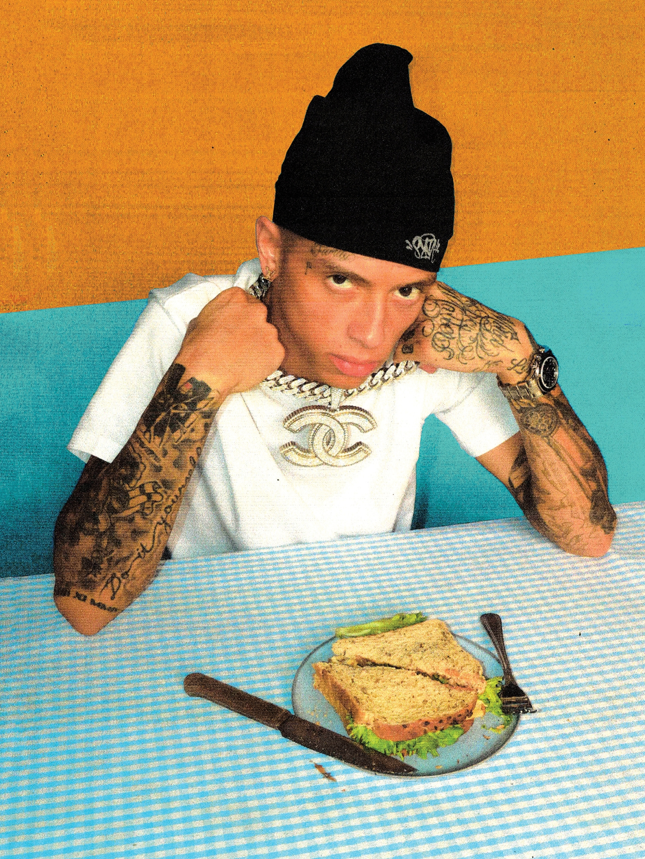 Adult male with tattoos wearing a black beanie and jewelry at a table with a sandwich and knife indoors in a lifestyle shot.