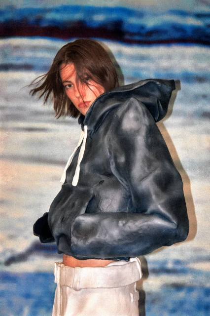 Adult female wearing a dark grey hoodie and light-colored pants poses in front of an abstract painted background with blue and white hues.