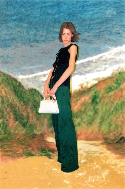 Fashion portrait of a young woman in a black top and green pants holding a white handbag, posing in front of a landscape backdrop.