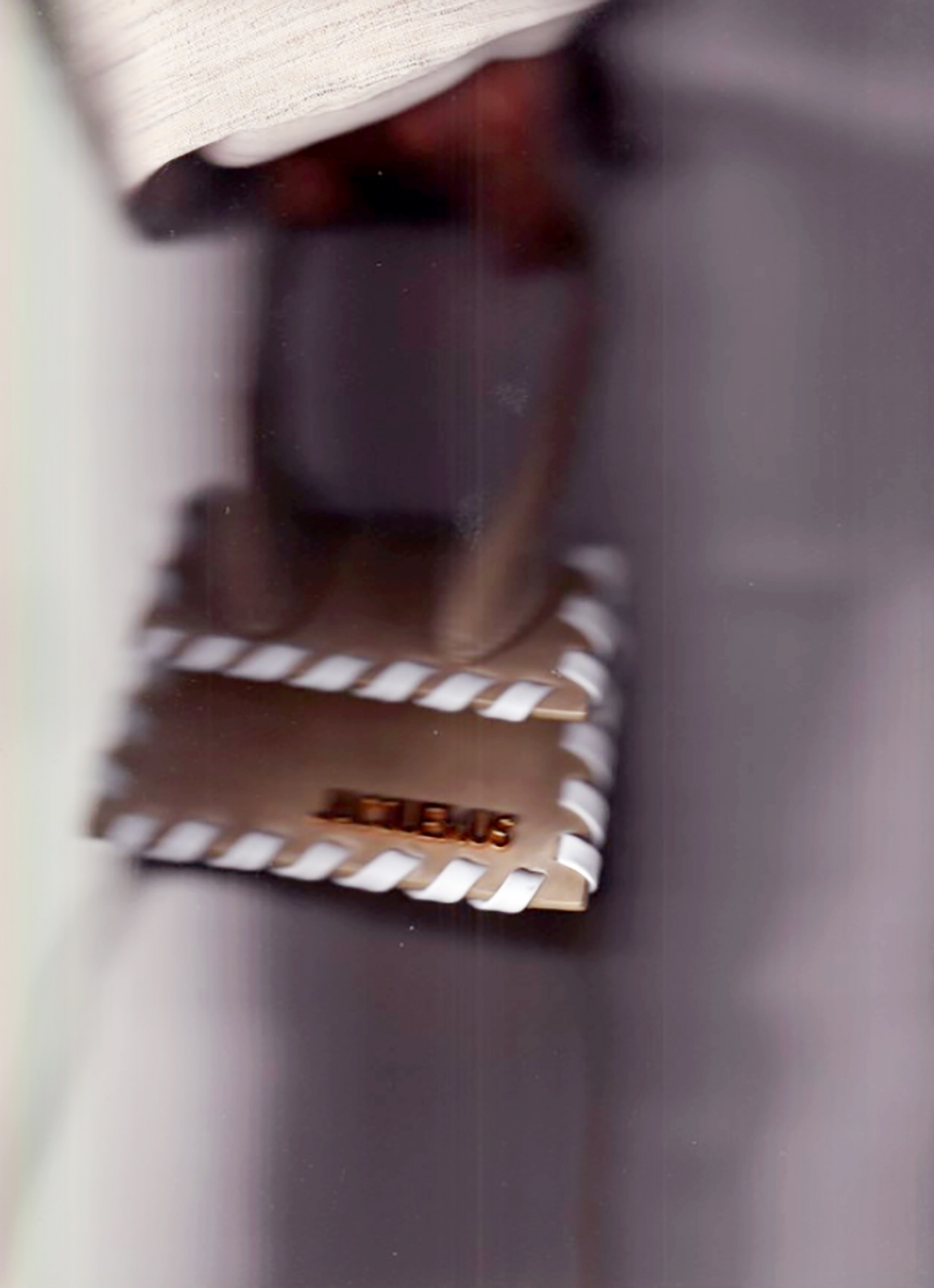 A tan Jacquemus handbag with white trim held by a person, seen from a close angle in a blurred indoor setting.