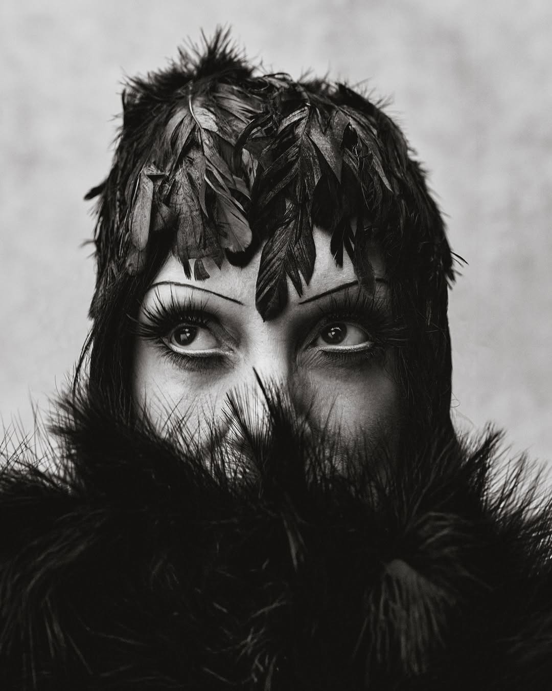Striking black and white portrait of a person with dramatic makeup, feathered headpiece, and fur collar, eyes looking upward.