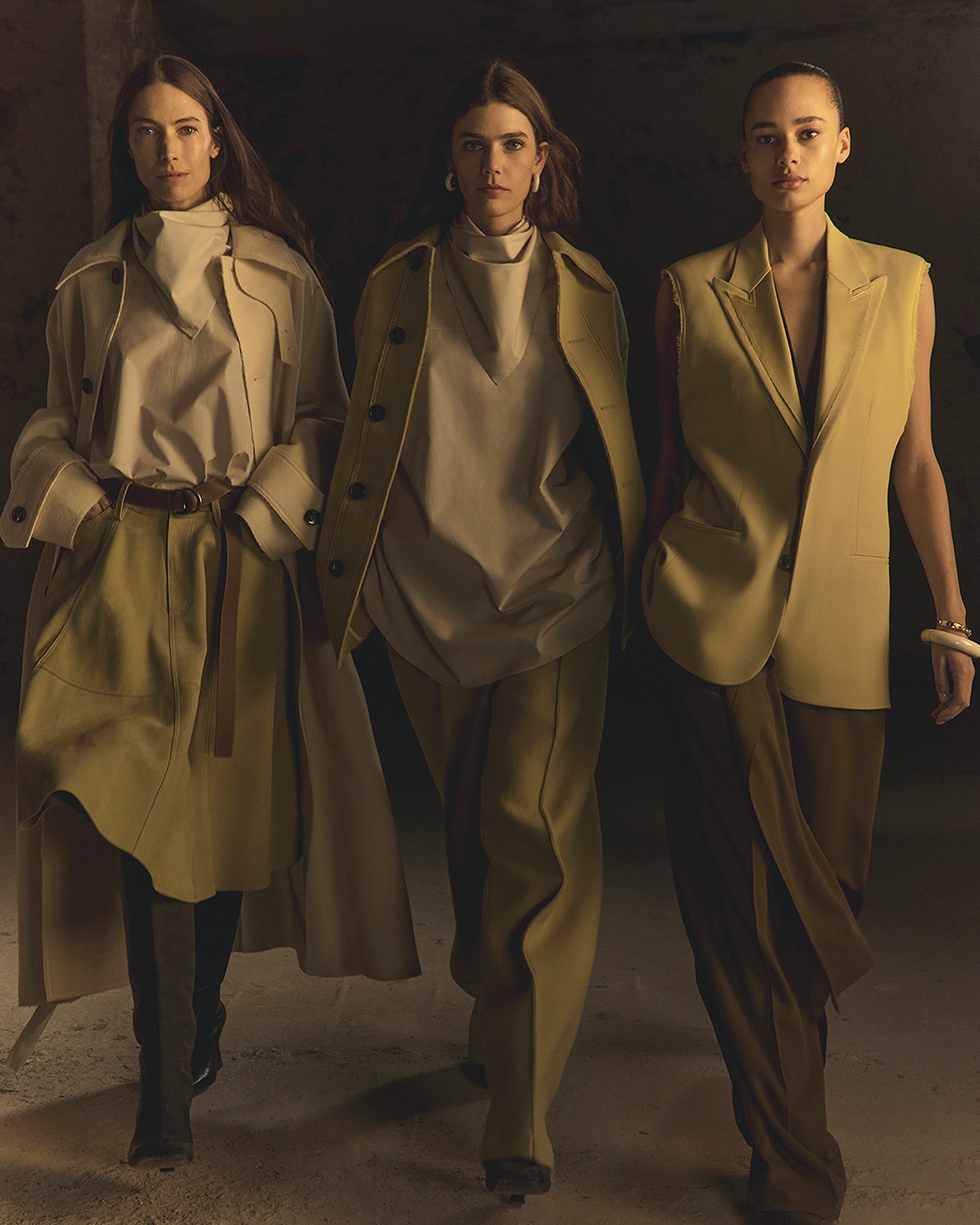 Three fashion models in beige outfits walk indoors; one wears a long coat, another a vest, photographed with neutral color grading.