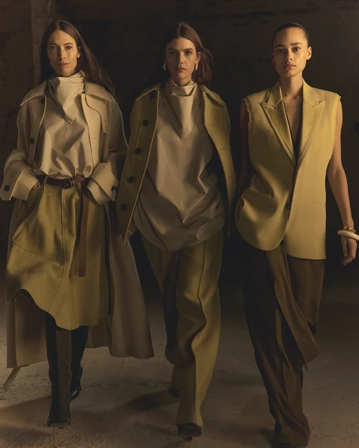 Three fashion models in beige outfits walk indoors; one wears a long coat, another a vest, photographed with neutral color grading.