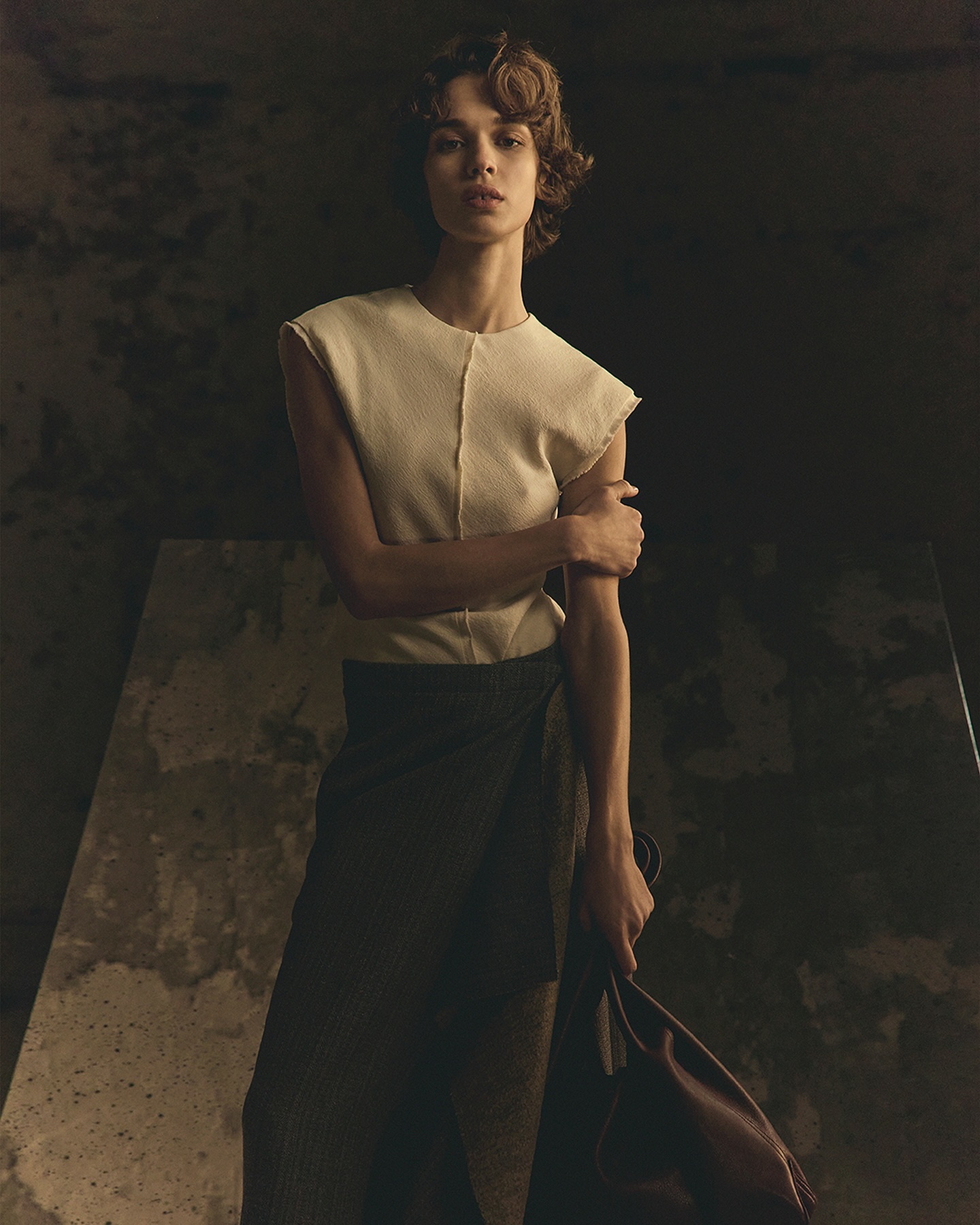 Elegant woman poses in studio, wearing cream top and dark wrap skirt, holding brown bag, with soft lighting and dark backdrop.