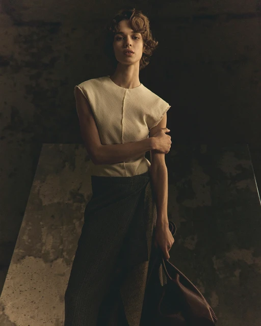 Elegant woman poses in studio, wearing cream top and dark wrap skirt, holding brown bag, with soft lighting and dark backdrop.