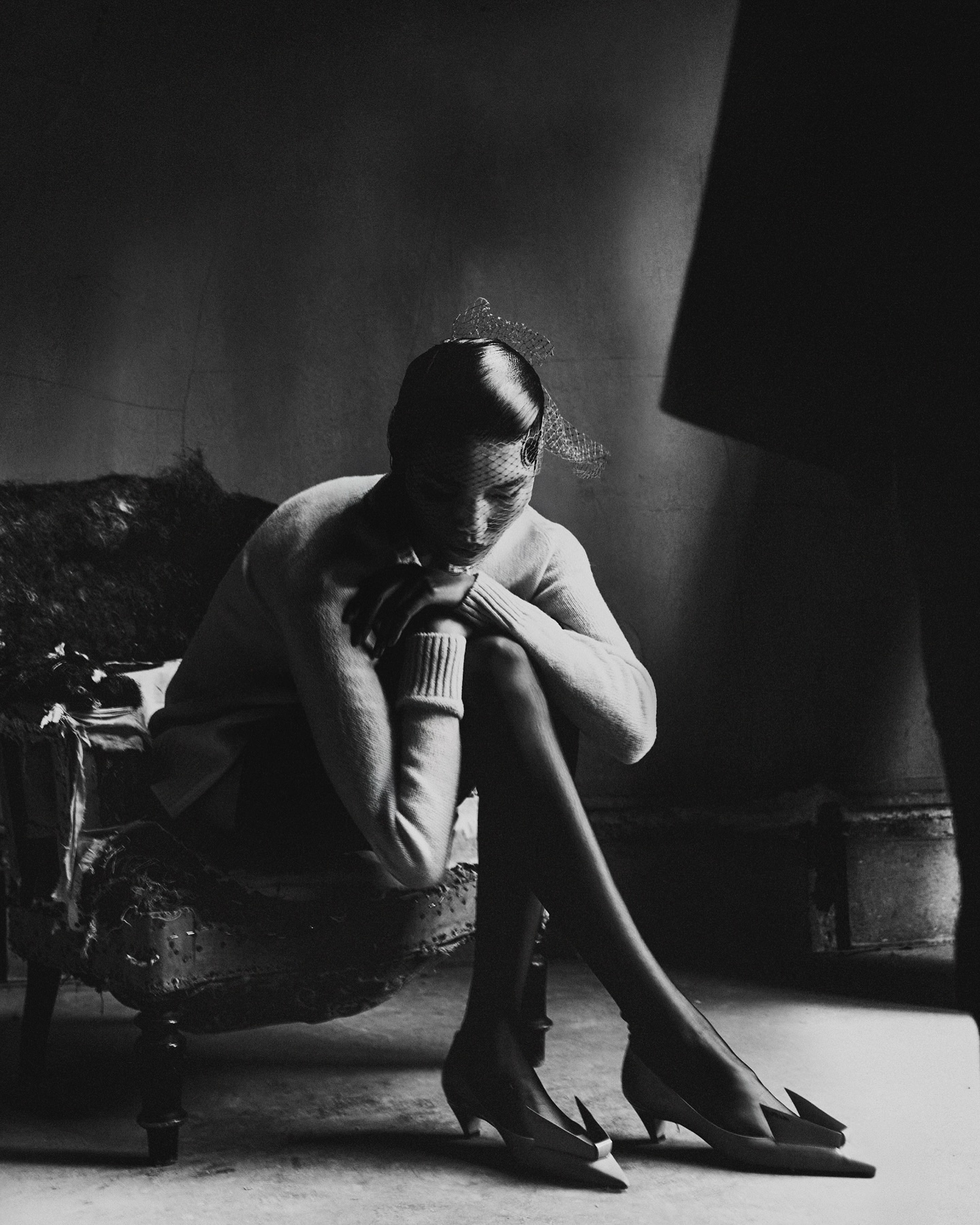 Elegant woman in pointed heels poses while seated in an upholstered chair, set against a subdued gray backdrop with diffused lighting.