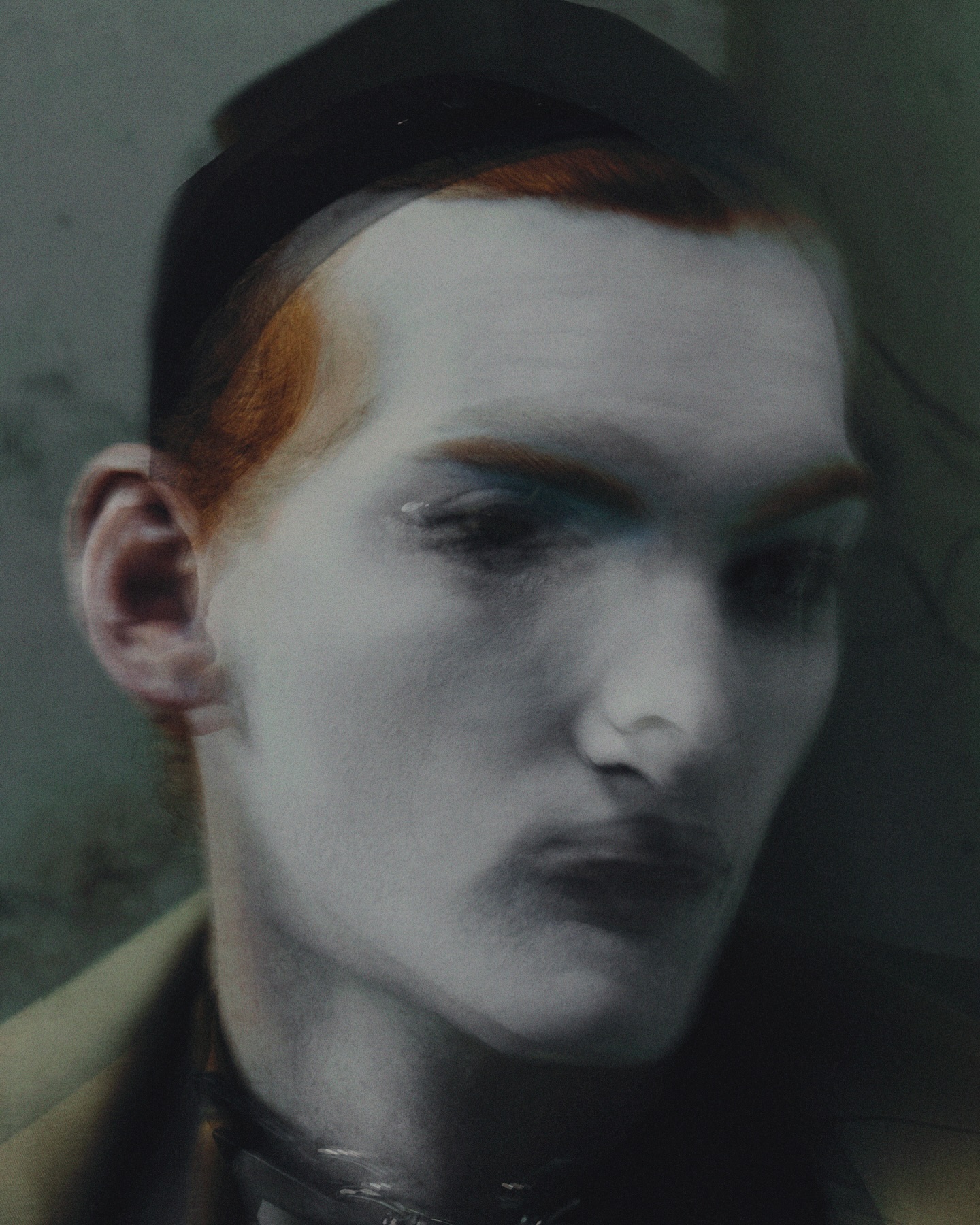 Ethereal beauty shot of an adult with pale skin, red hair, wearing a black hat and choker-style necklace against a hazy, grey-green backdrop.