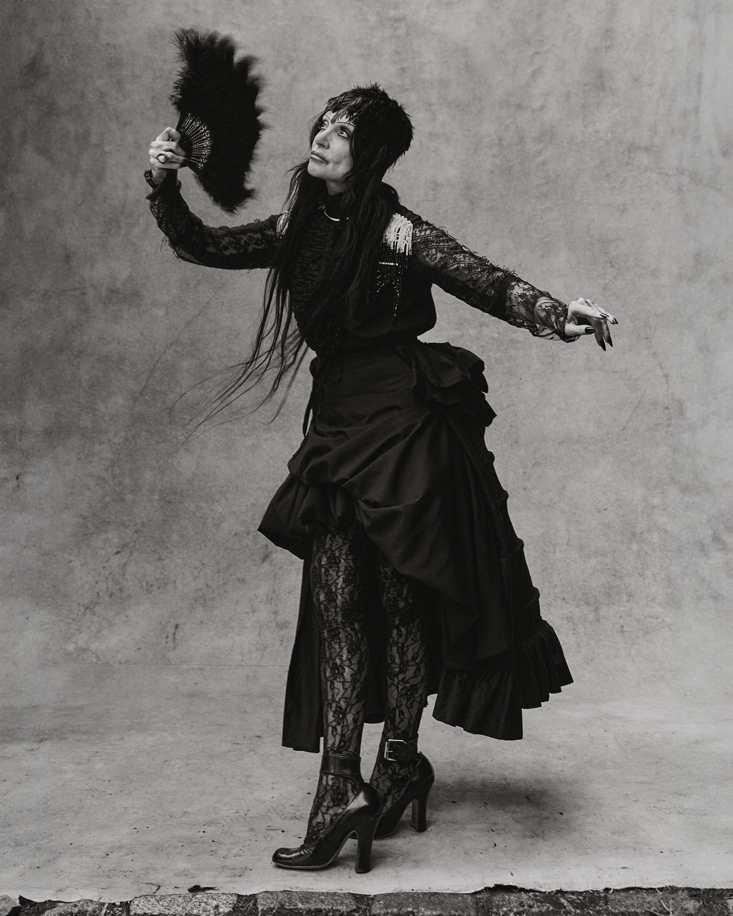 Monochrome full shot of an adult woman in a black dress with long dark hair, holding a feathered fan and looking up in a studio.