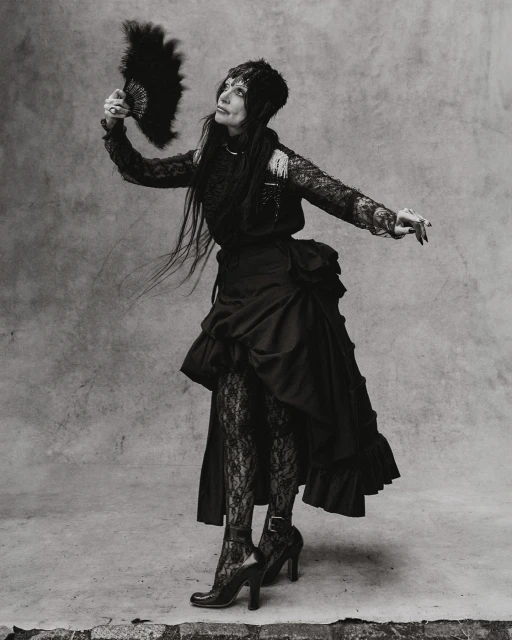 Monochrome full shot of an adult woman in a black dress with long dark hair, holding a feathered fan and looking up in a studio.