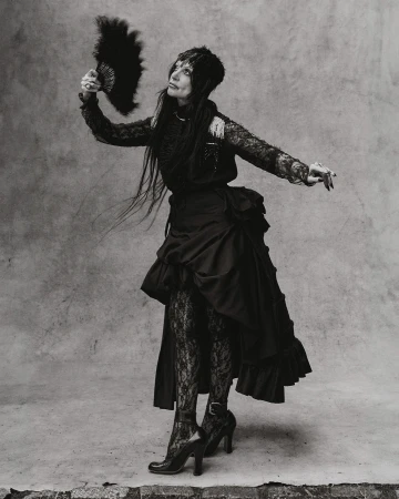Monochrome full shot of an adult woman in a black dress with long dark hair, holding a feathered fan and looking up in a studio.