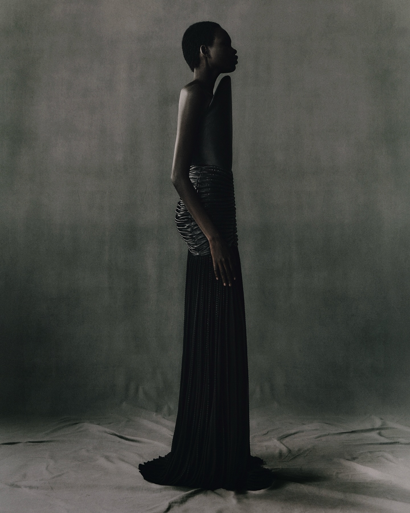 Profile shot of an adult woman with short hair wearing a black dress and cage-like corset in a studio with a grey backdrop.
