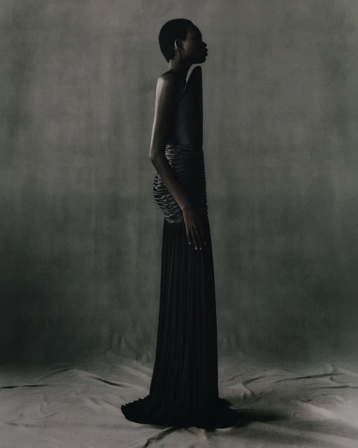 Profile shot of an adult woman with short hair wearing a black dress and cage-like corset in a studio with a grey backdrop.
