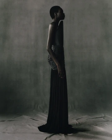 Profile shot of an adult woman with short hair wearing a black dress and cage-like corset in a studio with a grey backdrop.