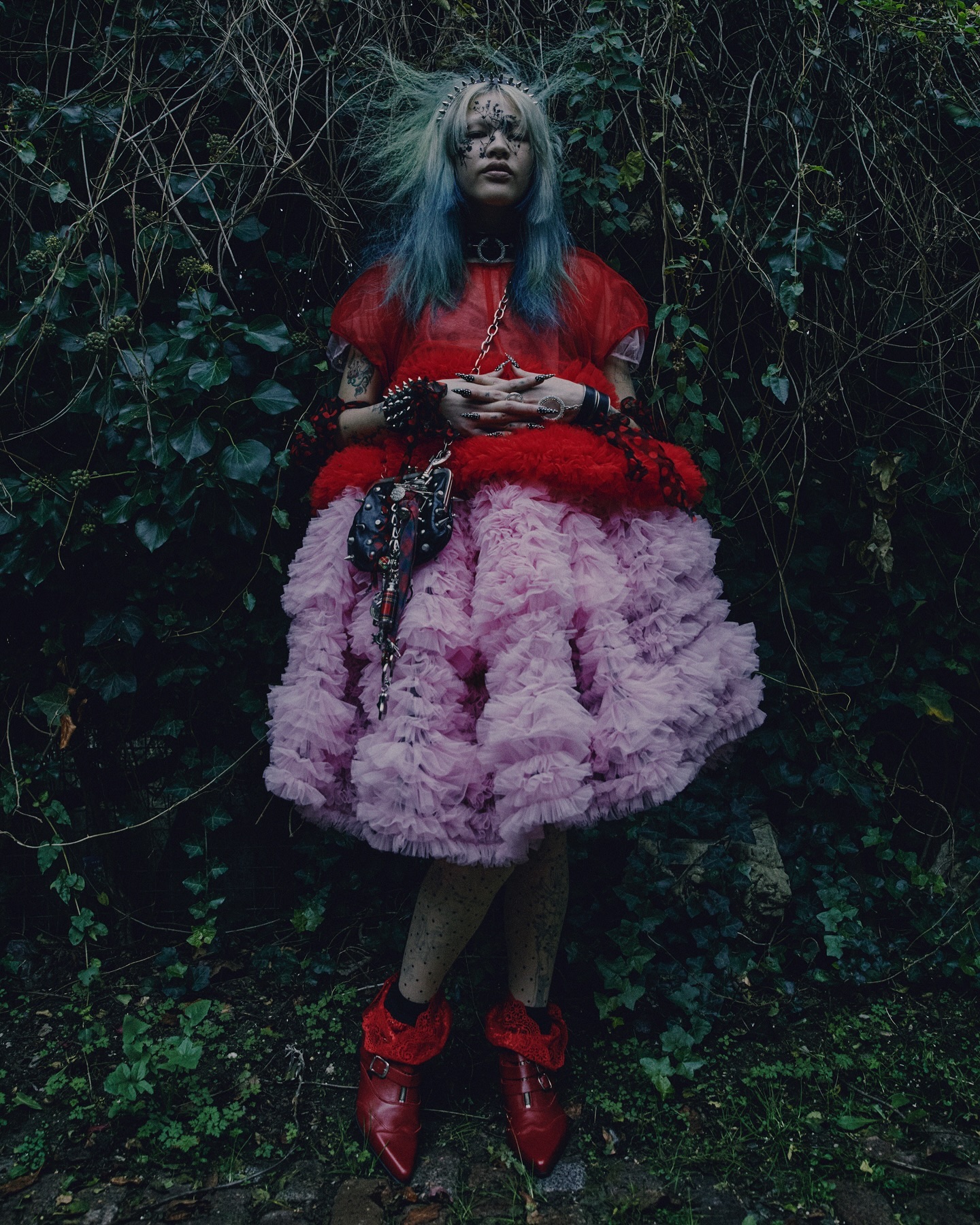 A single person with blue-green hair wears a red top and pink ruffled skirt outdoors surrounded by foliage, with red shoes and a purse.