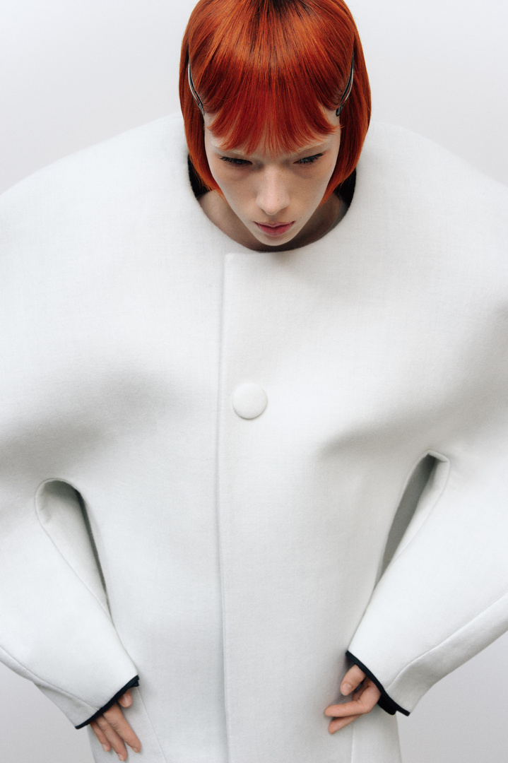 A red-haired woman poses indoors, wearing a sculptural white coat with a button, against a plain backdrop in a fashion setting.