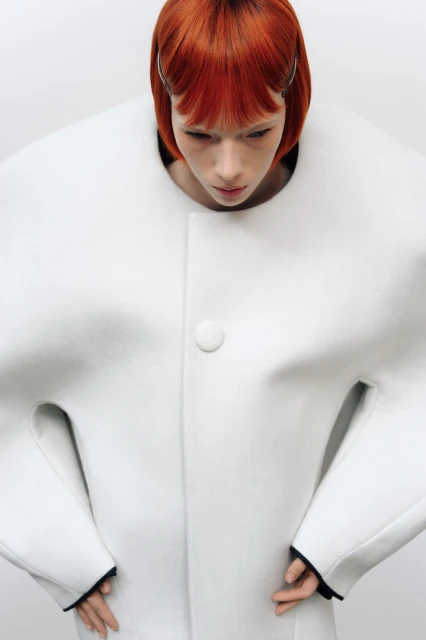 A red-haired woman poses indoors, wearing a sculptural white coat with a button, against a plain backdrop in a fashion setting.