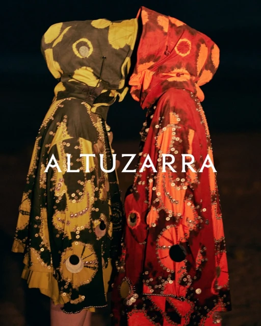 Two people in vibrant, patterned hooded cloaks, face-to-face in dim lighting. Decorative embellishments and the 'Altuzarra' logo are visible.
