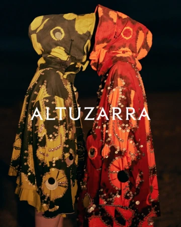 Two people in vibrant, patterned hooded cloaks, face-to-face in dim lighting. Decorative embellishments and the 'Altuzarra' logo are visible.