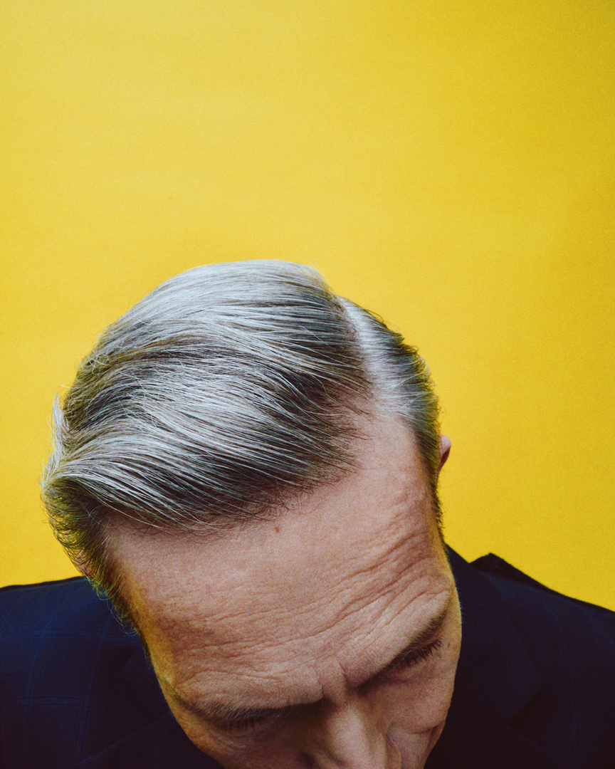 Close-up studio shot of a Caucasian man in a suit with grey hair against a bright yellow backdrop, capturing a sophisticated and stylish aesthetic.