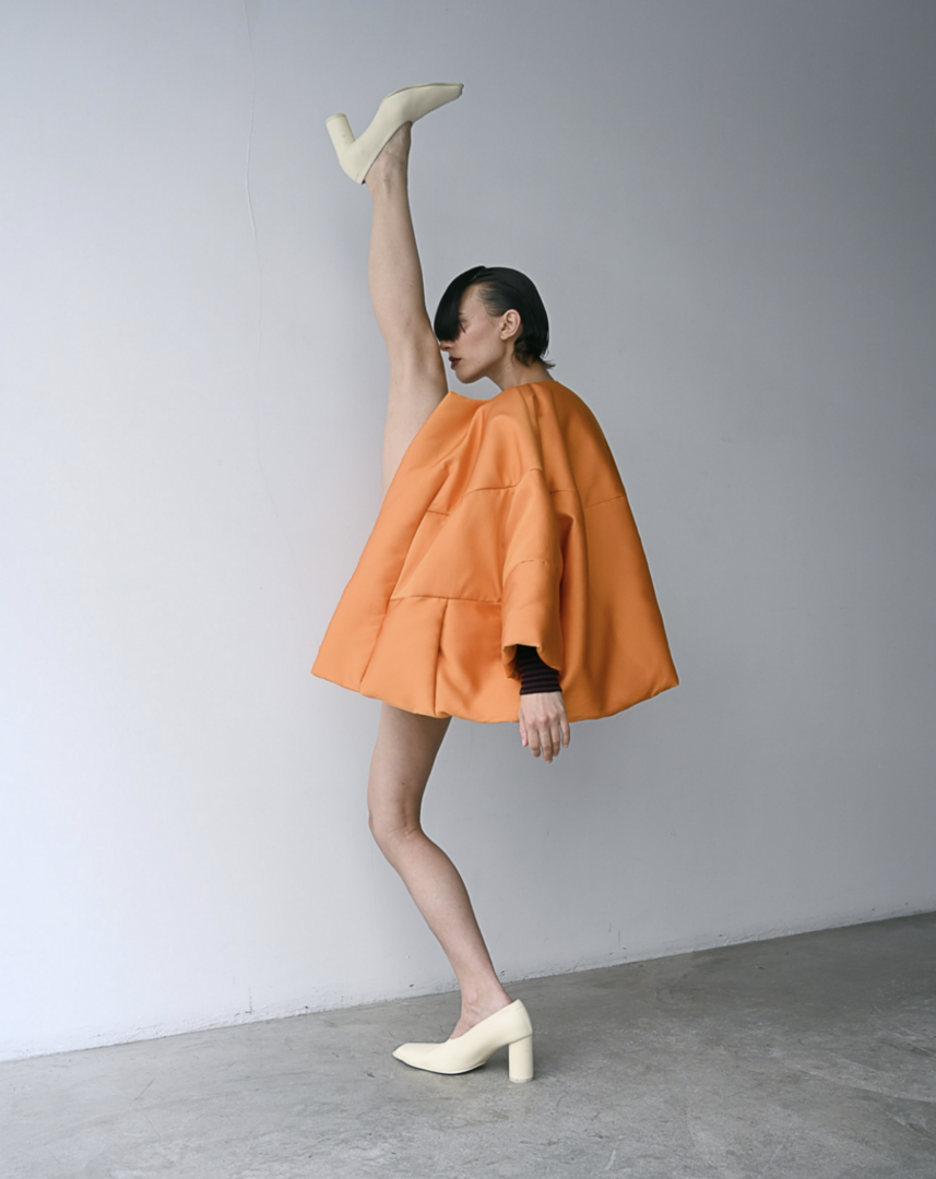 Fashionable woman in orange coat and pale yellow high heels posing in front of white wall, one leg extended upwards in vertical split.