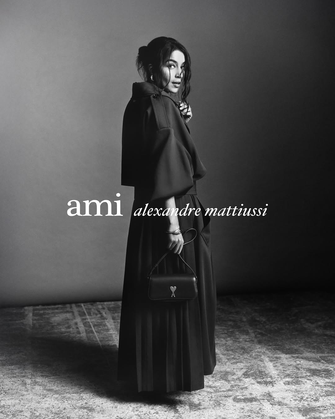 Black and white fashion portrait of a woman wearing a dark coat and long skirt holding a small black purse against a gray backdrop.