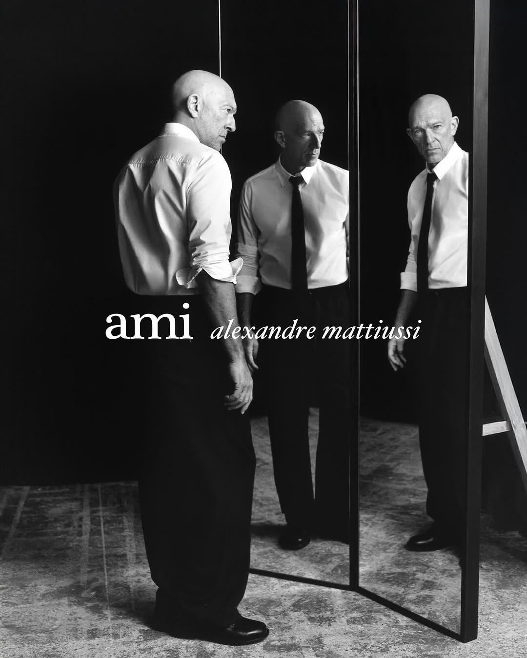 Black and white portrait of a bald adult male posing in front of a mirror wearing a shirt and tie with the 'ami' logo.