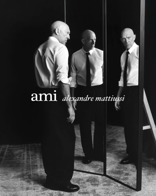 Black and white portrait of a bald adult male posing in front of a mirror wearing a shirt and tie with the 'ami' logo.