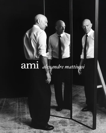 Black and white portrait of a bald adult male posing in front of a mirror wearing a shirt and tie with the 'ami' logo.