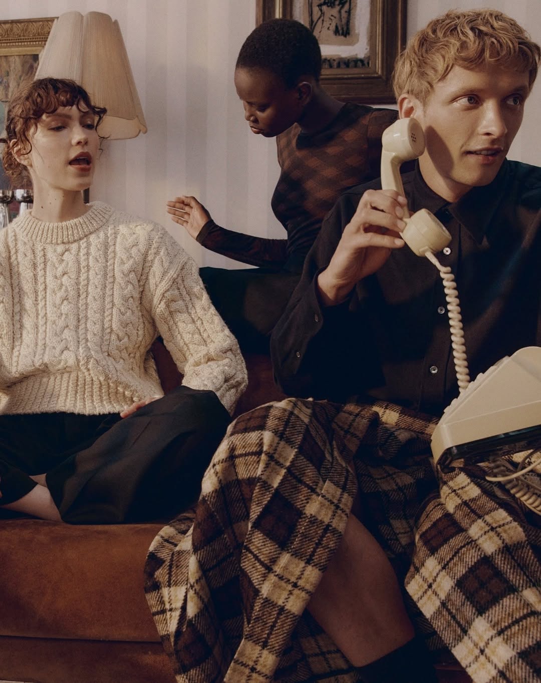 Three models in an indoor setting. A woman with short curly hair wears an ivory cable-knit sweater, and a man in a plaid skirt holds a vintage telephone.