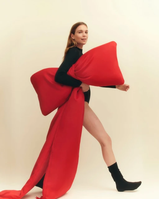 Woman in black bodysuit walks with large red pillow with attached flowing red fabric, set against a neutral off-white studio backdrop.