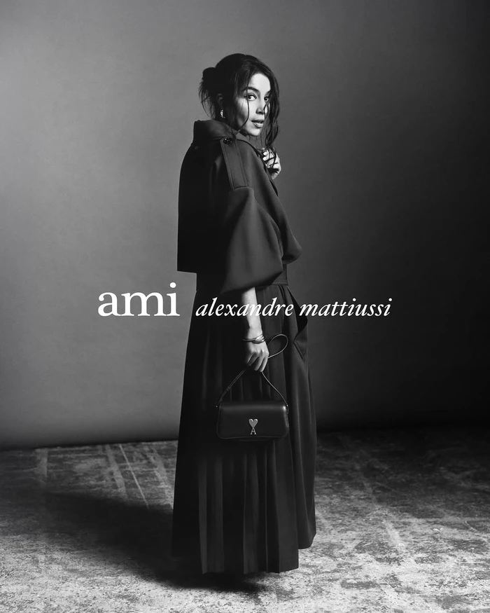Monochrome fashion portrait of a woman in a dark belted coat holding a small handbag against a plain studio background.