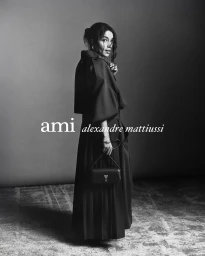 Monochrome fashion portrait of a woman in a dark belted coat holding a small handbag against a plain studio background.