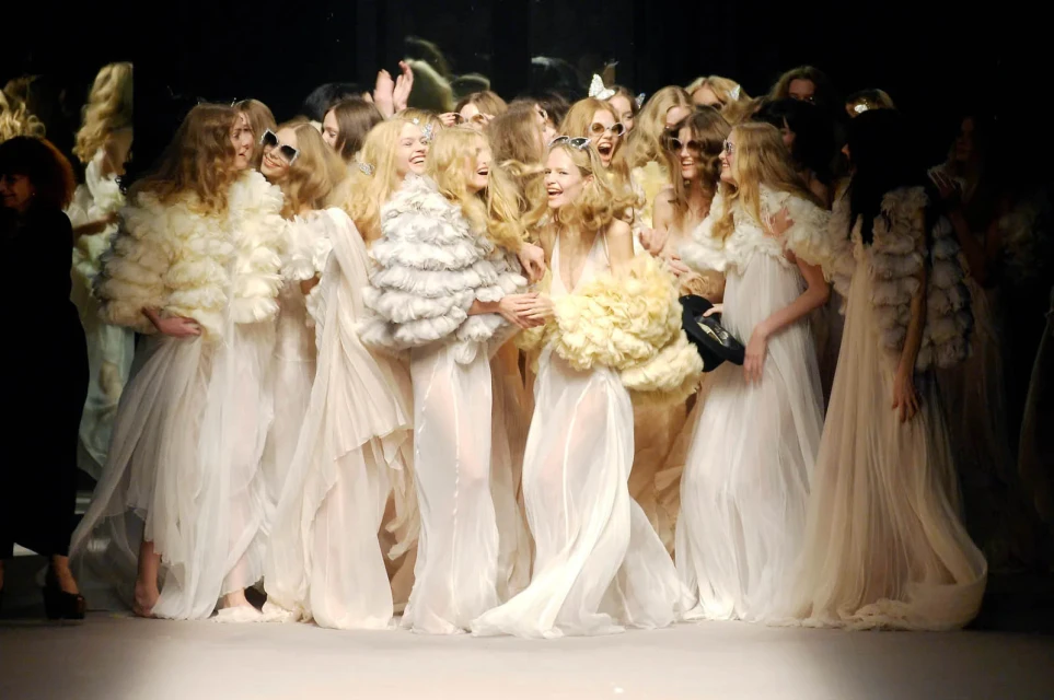 A group of fashion models on a runway show, dressed in sheer dresses and ruffled boleros. They are laughing and posing together.
