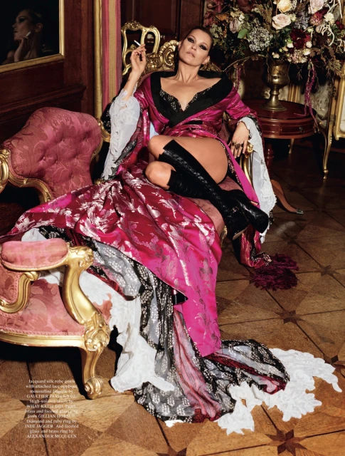 Fashion shot of an adult female in a patterned pink robe, black boots, and jewelry sitting on an ornate chair in an indoor setting.