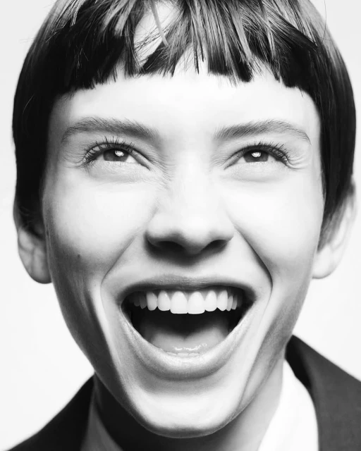 Black and white portrait of a young woman with a cropped haircut smiling broadly with an open mouth and visible teeth indoors.