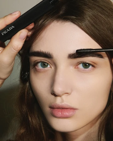 A woman's face in a beauty shot with a makeup artist applying eyebrow gel with a brush and holding a Prada product. Close-up.