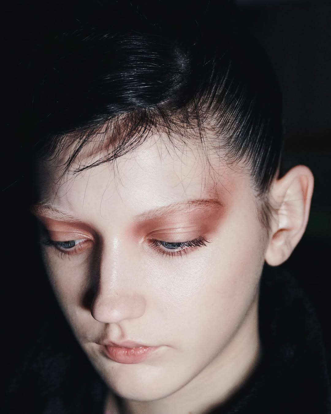 Close-up portrait of a young, light-skinned person with dark hair and striking eyeshadow, looking down in a dimly lit setting.