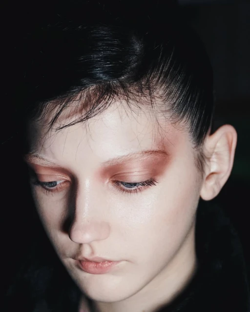 Close-up portrait of a young, light-skinned person with dark hair and striking eyeshadow, looking down in a dimly lit setting.