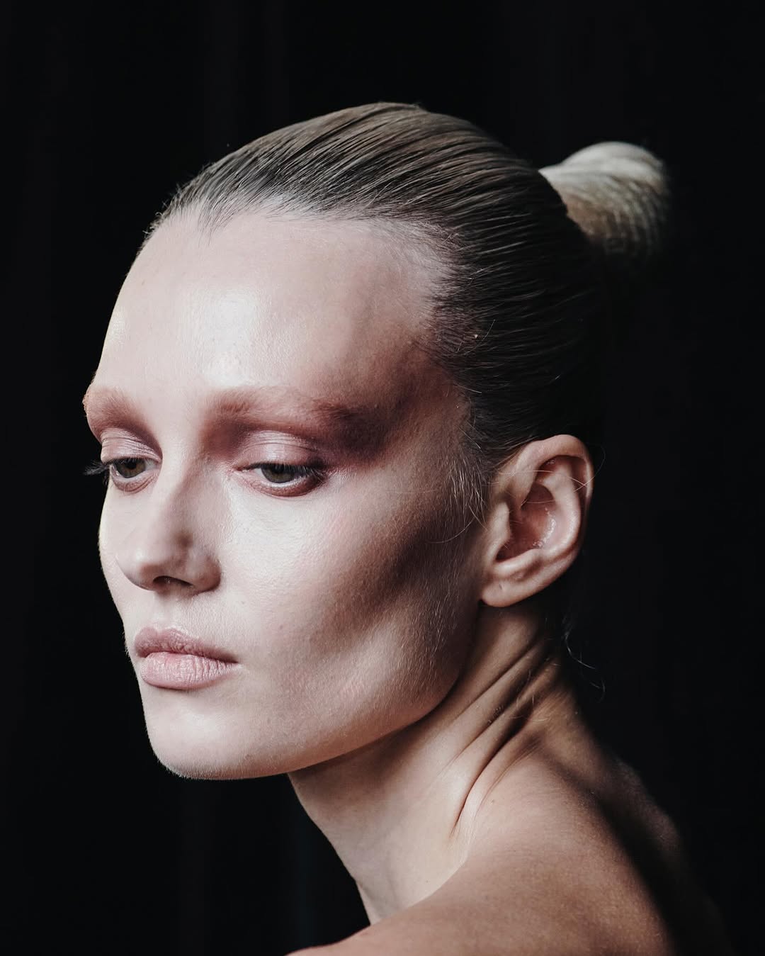 Portrait of adult woman with light skin, with slicked-back hair in a bun and wearing subtle makeup, against a dark background.