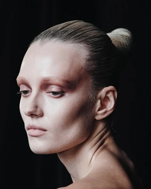 Portrait of adult woman with light skin, with slicked-back hair in a bun and wearing subtle makeup, against a dark background.