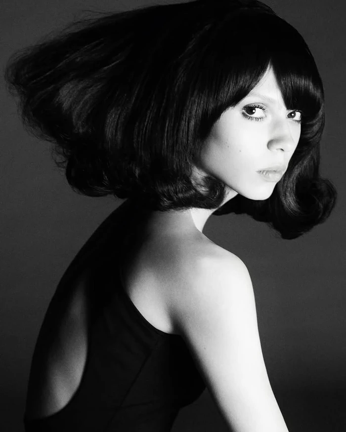 Black and white portrait of a woman with voluminous dark bob hair looking over her shoulder against dark background.