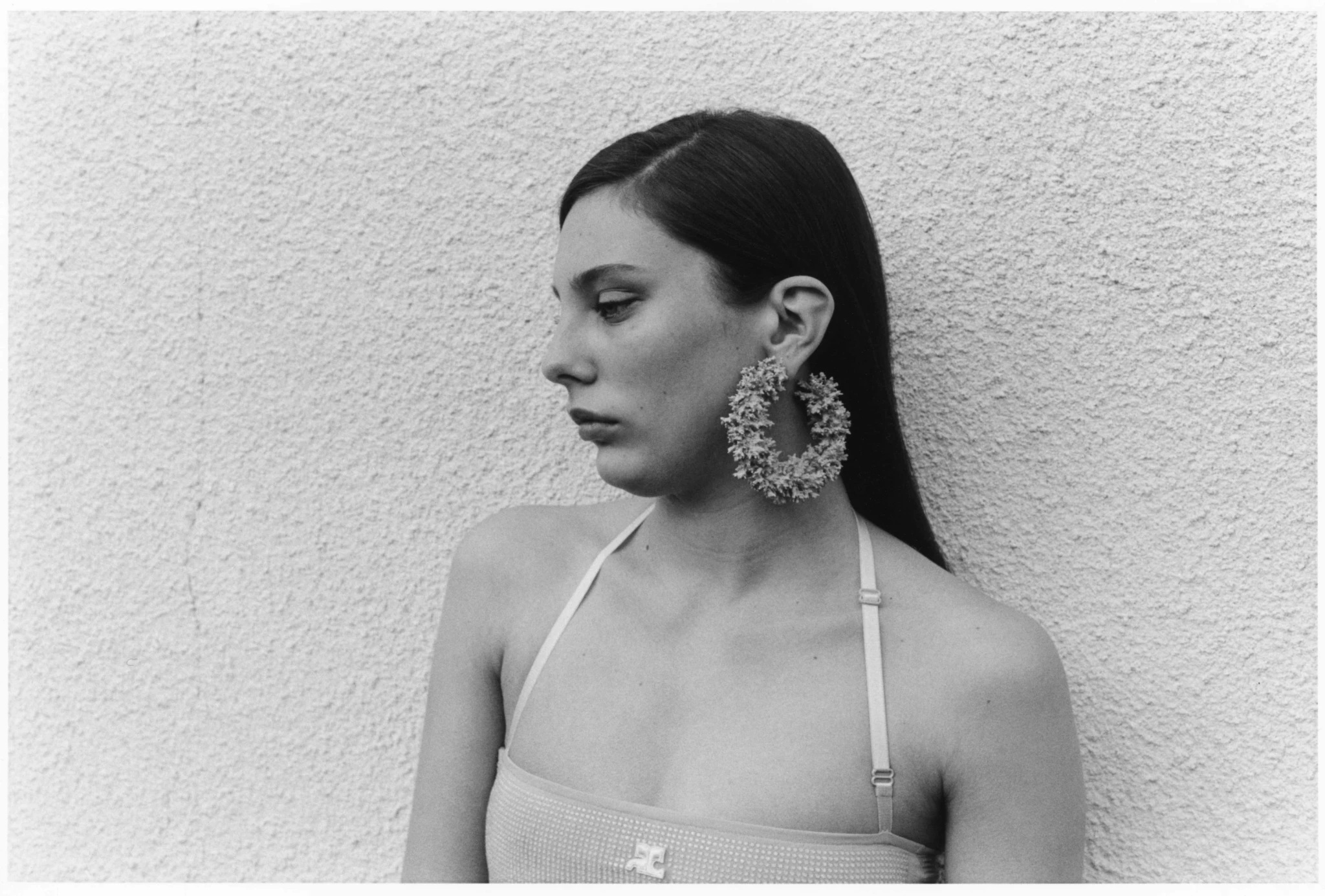 Portrait of a woman in profile wearing floral hoop earrings, a spaghetti-strap top, and dark hair, posing in front of a textured wall.
