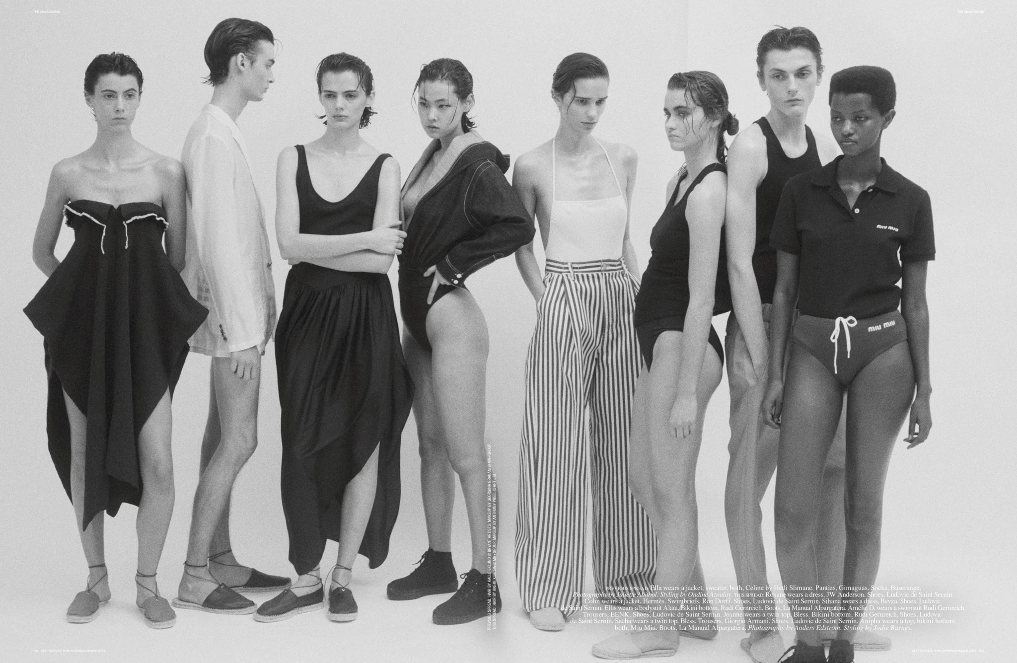 Black-and-white fashion editorial featuring eight diverse models in designer clothing posing together in a photo studio on a seamless white background.