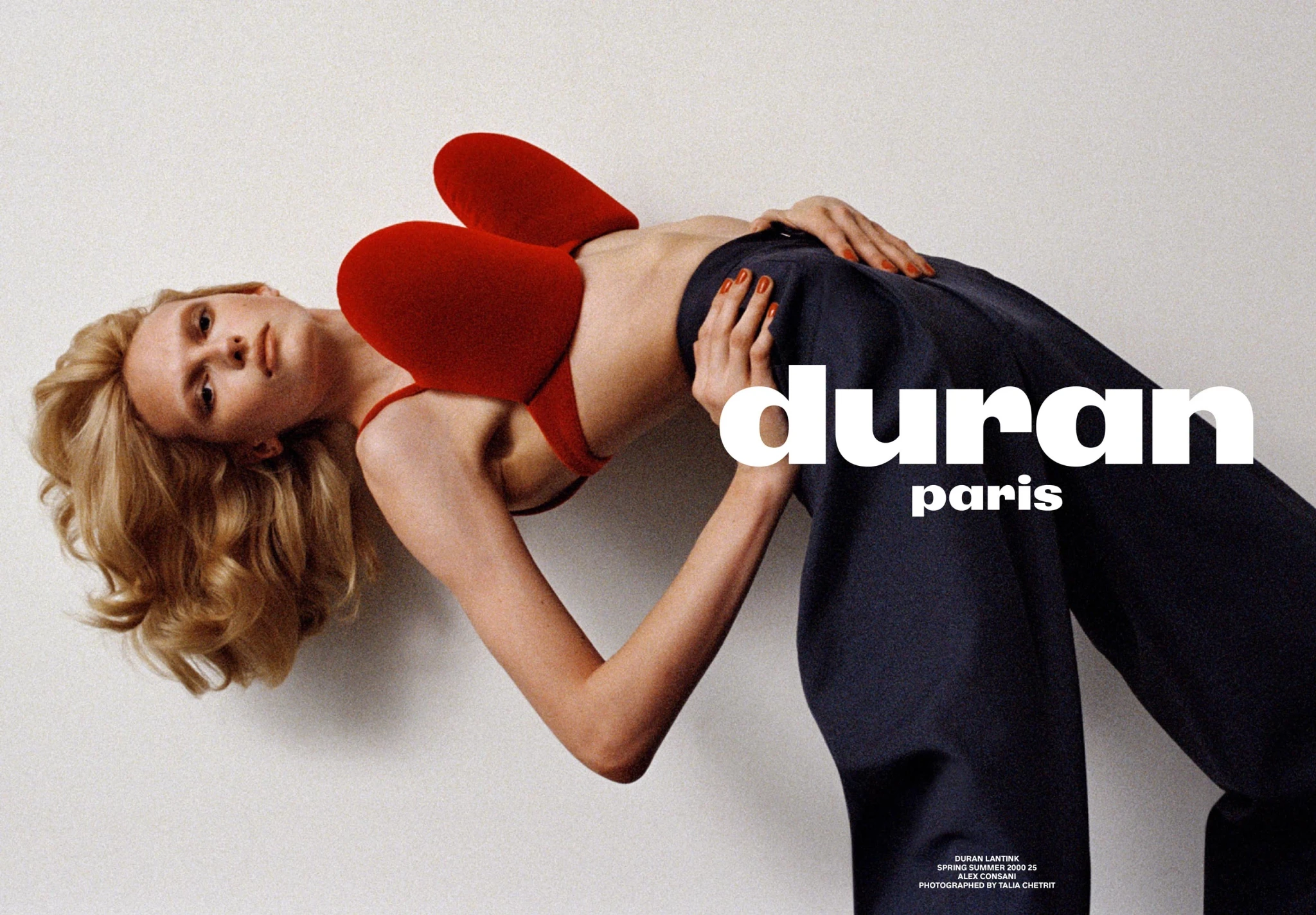 Fashion editorial shot of a blonde woman in a red bra and dark blue trousers leaning back, with studio lighting and a plain white backdrop.